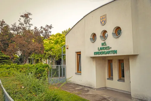 Ein Kindergartengebäude mit einem Schild, das 'Nr. Landes-Kindergarten' über dem Eingang zeigt, mit drei runden Fenstern und einem kleinen Wappen über dem Schild.