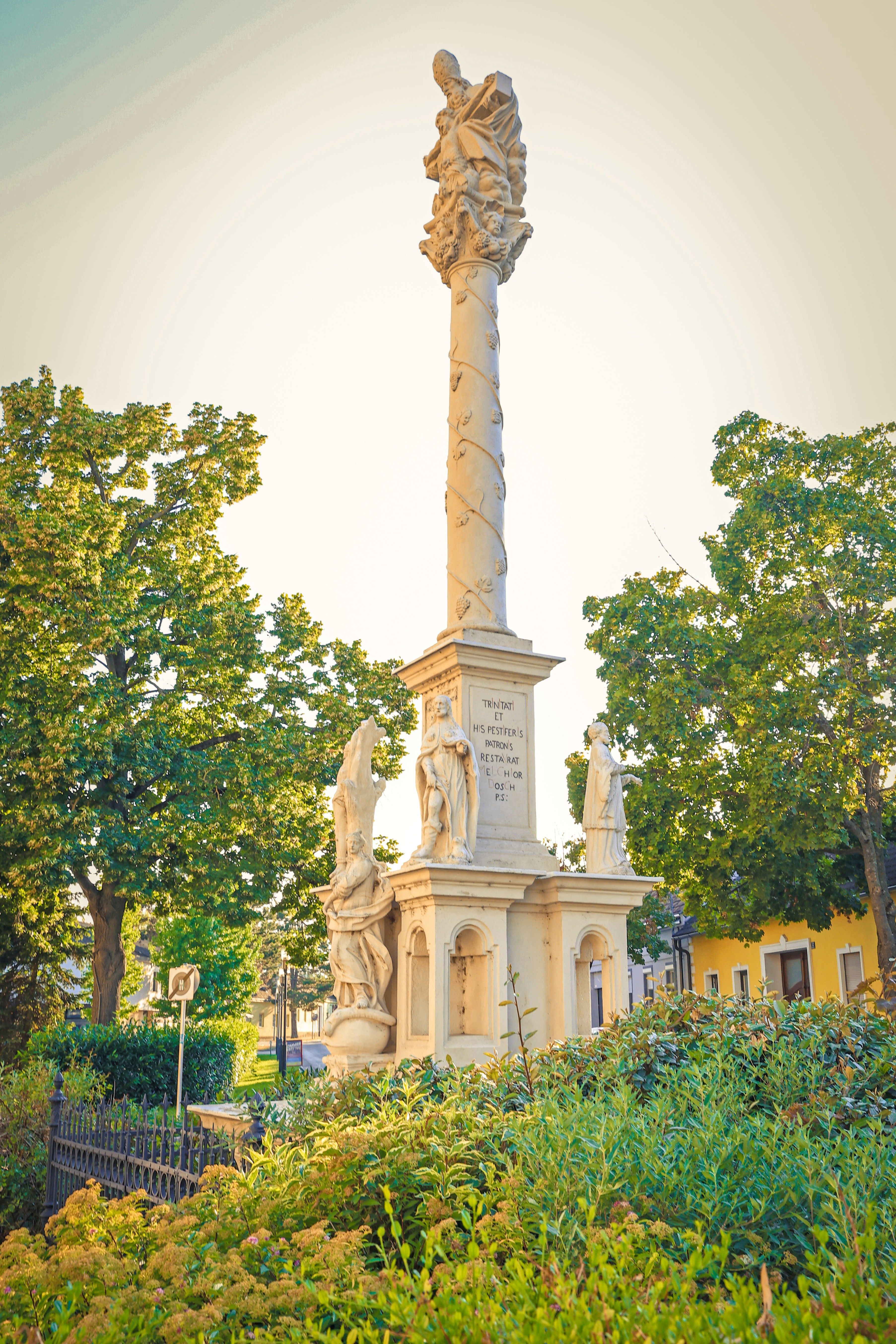 Ein hoher, kunstvoll verzierter Steinmonument steht in einem Park, umgeben von Bäumen und Grün. Es zeigt Statuen von drei Figuren mit einer Inschrift an seiner Basis. Der Hintergrund des Monuments ist ein gelbes Gebäude.