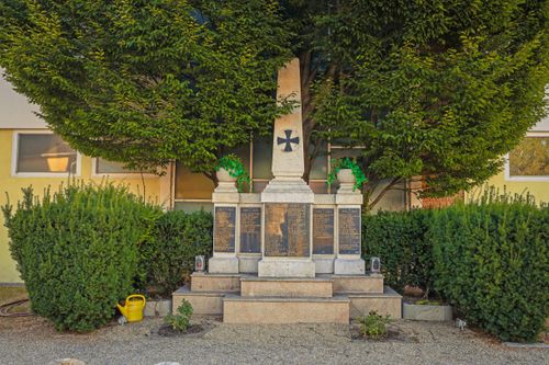 Ein Denkmal mit einem Kreuz oben, umgeben von Pflanzen und Büschen, mit auf den Seiten eingravierten Tafeln.