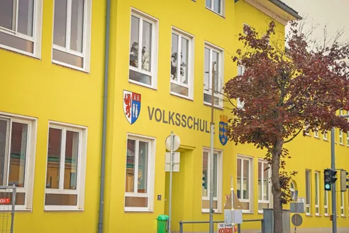 Ein gelbes Gebäude mit weißen Fenstern und einem Schild, das 'Volksschule' mit zwei Wappen darüber liest.