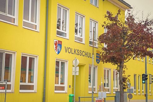 Ein gelbes Gebäude mit weißen Fenstern und einem Schild, das 'Volksschule' mit zwei Wappen darüber liest.