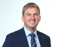 Ein Mann in einem dunkelblauen Anzug und einem blauen Hemd mit einem rot, weiß und blau gestreiften Krawatte lächelt und posiert für ein Foto.
