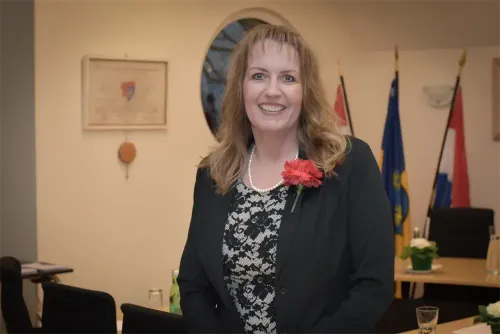 Eine Frau in einem schwarzen Blazer und einer Bluse mit Blumenmuster steht lächelnd. Sie hat eine rote Blume an ihrem Blazer und befindet sich in einem Raum mit Fahnen und einem Bilderrahmen.
