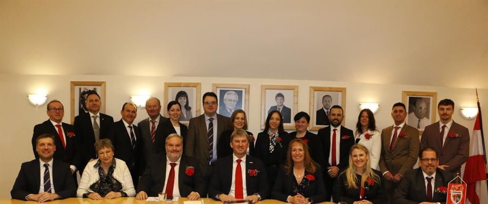 Eine Gruppe von Menschen in formeller Kleidung posiert für ein Foto in einem Konferenzraum mit gerahmten Porträts an der Wand.