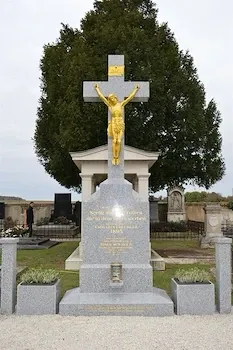 Ein goldenes Kruzifix steht prominent auf einem grauen Denkmal auf einem Friedhof, umgeben von Grün und einem großen Baum.