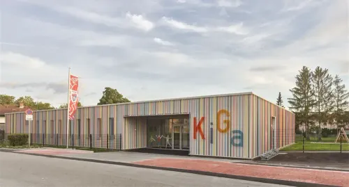 Ein farbenfrohes Gebäude mit großen Buchstaben, die 'Kiga' auf der Seite lesen, einem Fahnenmast und einem Bürgersteig.