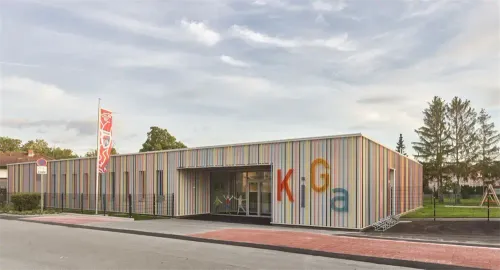 Ein farbenfrohes Gebäude mit großen Buchstaben, die 'Kiga' auf der Seite lesen, einem Fahnenmast und einem Bürgersteig.