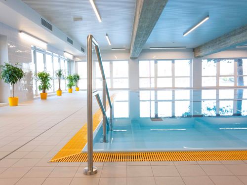 Ein Hallenbad mit einer gelben Sicherheitsspur, Edelstahl-Handläufen und mehreren Topfpflanzen in gelben Töpfen. Das Becken ist von großen Fenstern umgeben.