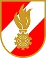 Das Emblem zeigt einen roten und weißen Schild mit einer goldenen Flamme in der Mitte, umgeben von einem Zahnrad-Design.