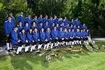 Eine große Gruppe von Personen in blauen Uniformen posiert für ein Foto auf einem Rasenfeld mit Musikinstrumenten und Bäumen im Hintergrund.