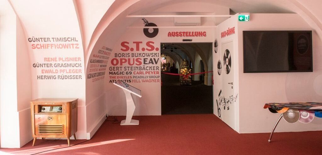 An exhibition space with a red carpet, featuring a wall with names and music notes, a sign reading 'S.T.S. OPUS EAV', and a display stand with a book.