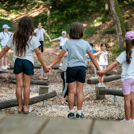 Eine Gruppe von Kindern spielt auf dem Spielplatz. Sie halten sich an den Händen und gehen auf den Holzhindernissen.