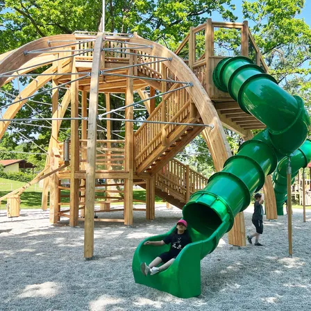 Kinder spielen auf einer hölzernen Spielhausstruktur mit einem grünen Spiralrutsche in einem Park. Andere Kinder sind im Hintergrund zu sehen.