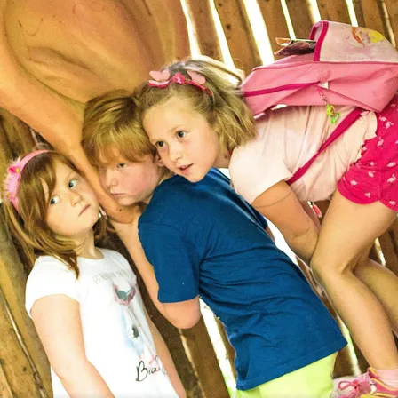 Drei Kinder stehen hinter einem Holzzaun, das Mädchen in Pink trägt einen Rucksack und der Junge in Blau trägt ein weißes Hemd.