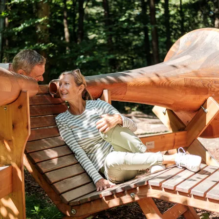 Ein älteres Paar sitzt auf einer Holzbank im Wald und schaut auf einen großen Holzhorn.