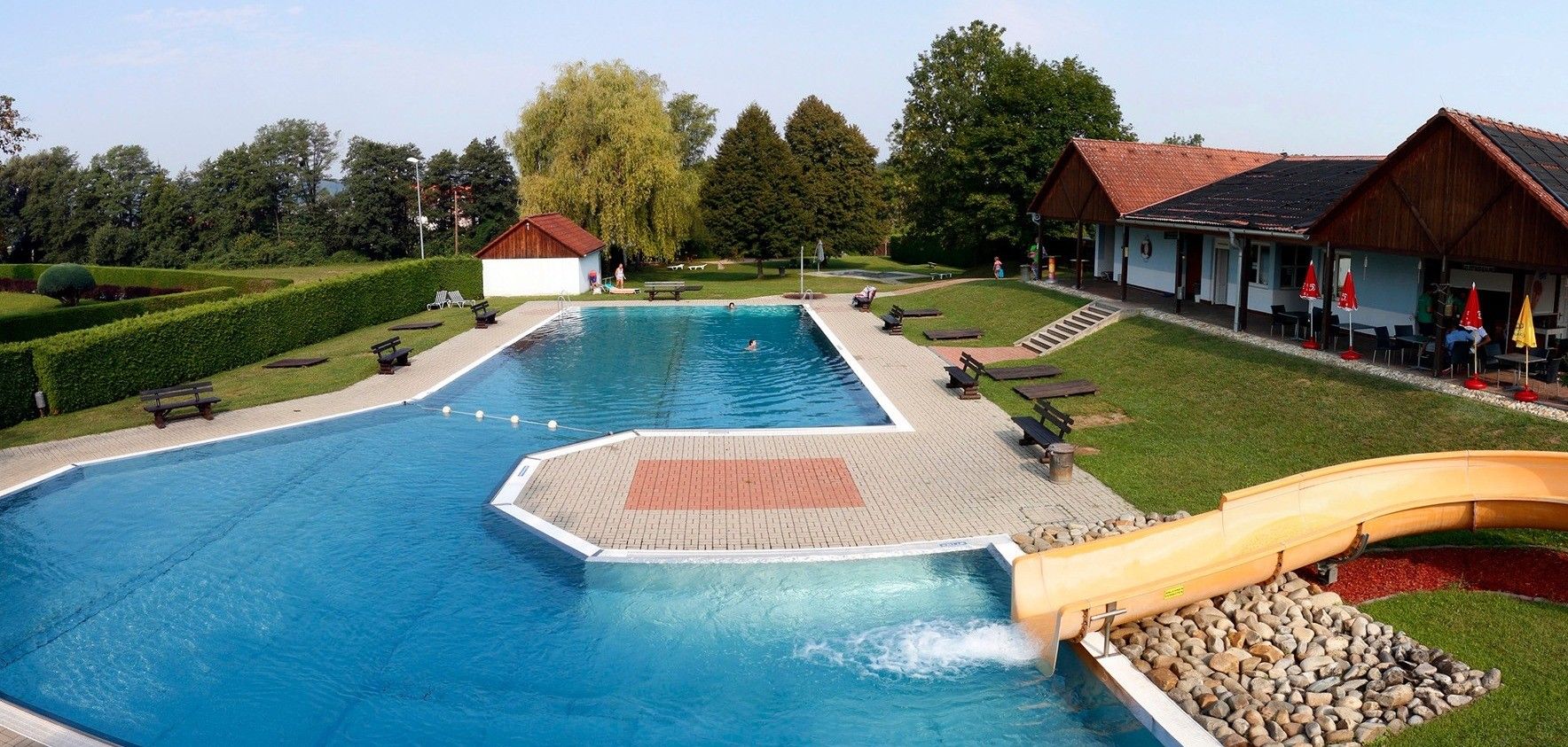Luftaufnahme eines großen Freibades, umgeben von viel Grün, mit zwei kleineren Becken, Bänken und einer Rutsche.