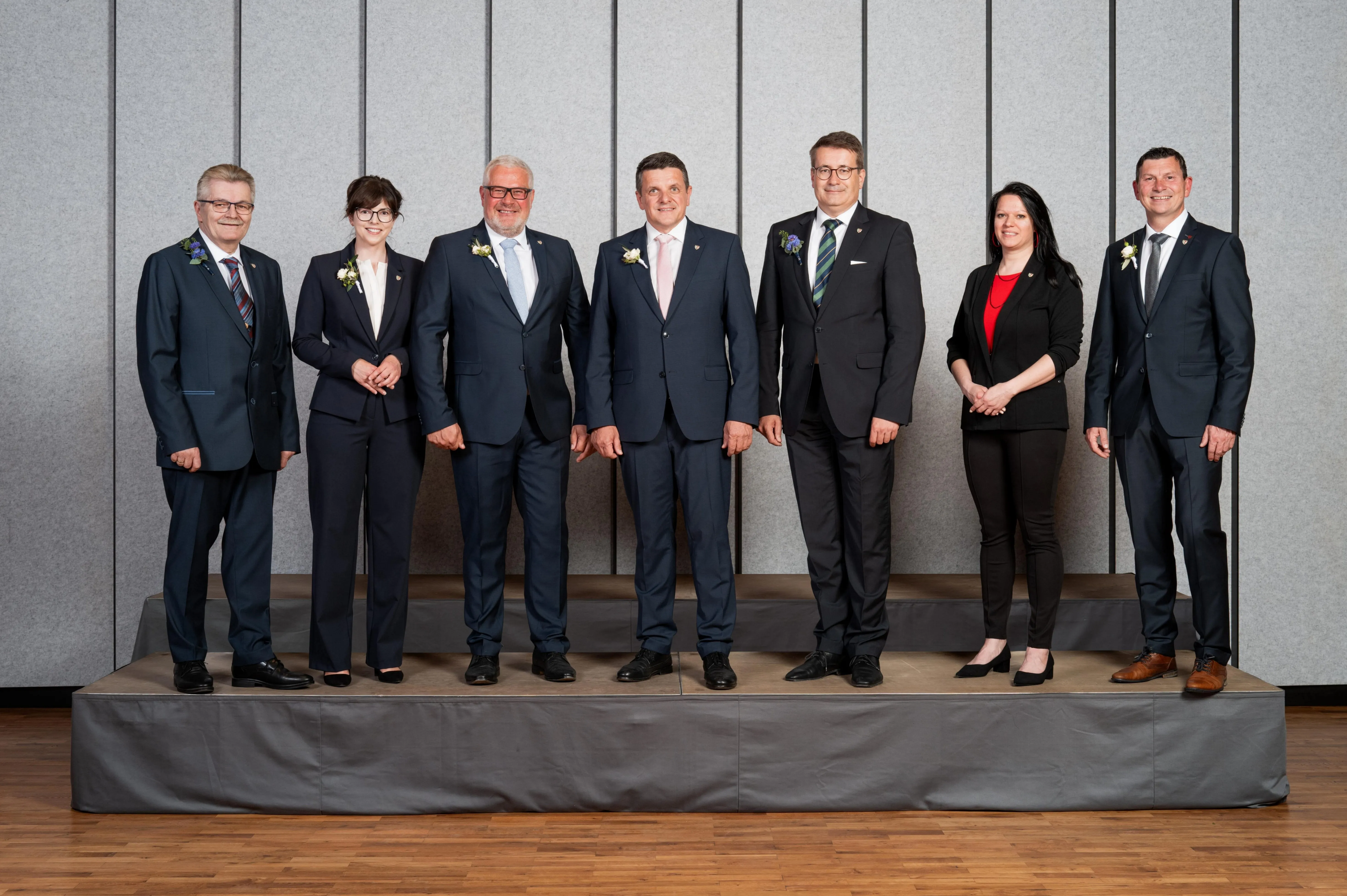 Eine Gruppe von Menschen in Anzügen steht auf einer Bühne vor einer grauen Wand und lächelt für ein Foto.