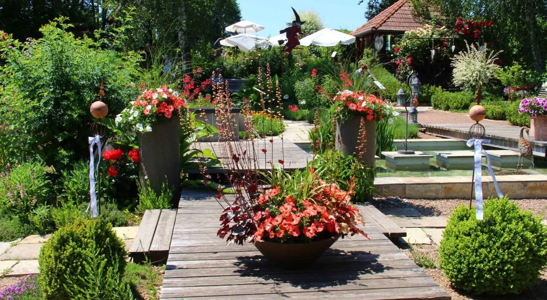 Ein Garten im Freien mit Holzterrasse, der einen großen runden Blumentopf mit roten und weißen Blumen zeigt, umgeben von üppigem Grün und einer Holzbank.