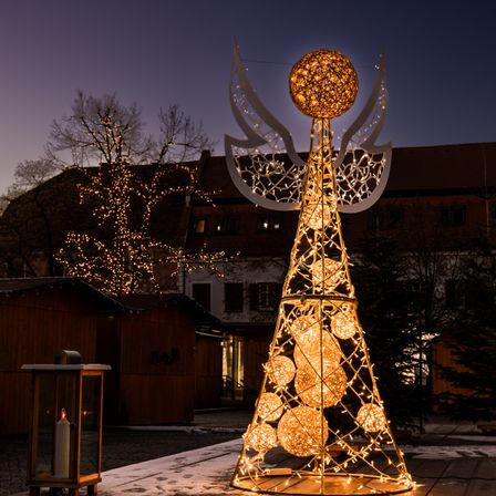 Ein Weihnachtsbaum mit einem goldenen Lichtdesign steht in einem Außenbereich. Der Baum ist von einem Zaun umgeben, und eine Kerzenlaterne steht links. Hinter dem Baum befinden sich einige Gebäude.