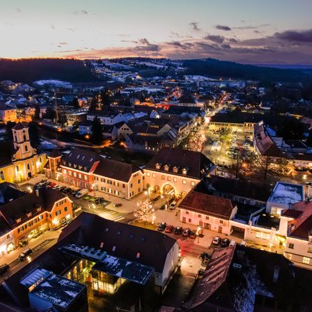 Eine Luftaufnahme einer Stadt mit vielen Gebäuden und Lichtern bei Nacht. Die Stadt ist von Bäumen und Bergen umgeben.