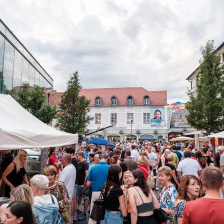 Ein überfüllter Straßenmarkt unter Regenschirmen mit Gebäuden im Hintergrund, eines mit einem großen Plakat. Menschen gehen herum, einige tragen Hüte und haben Taschen bei sich.
