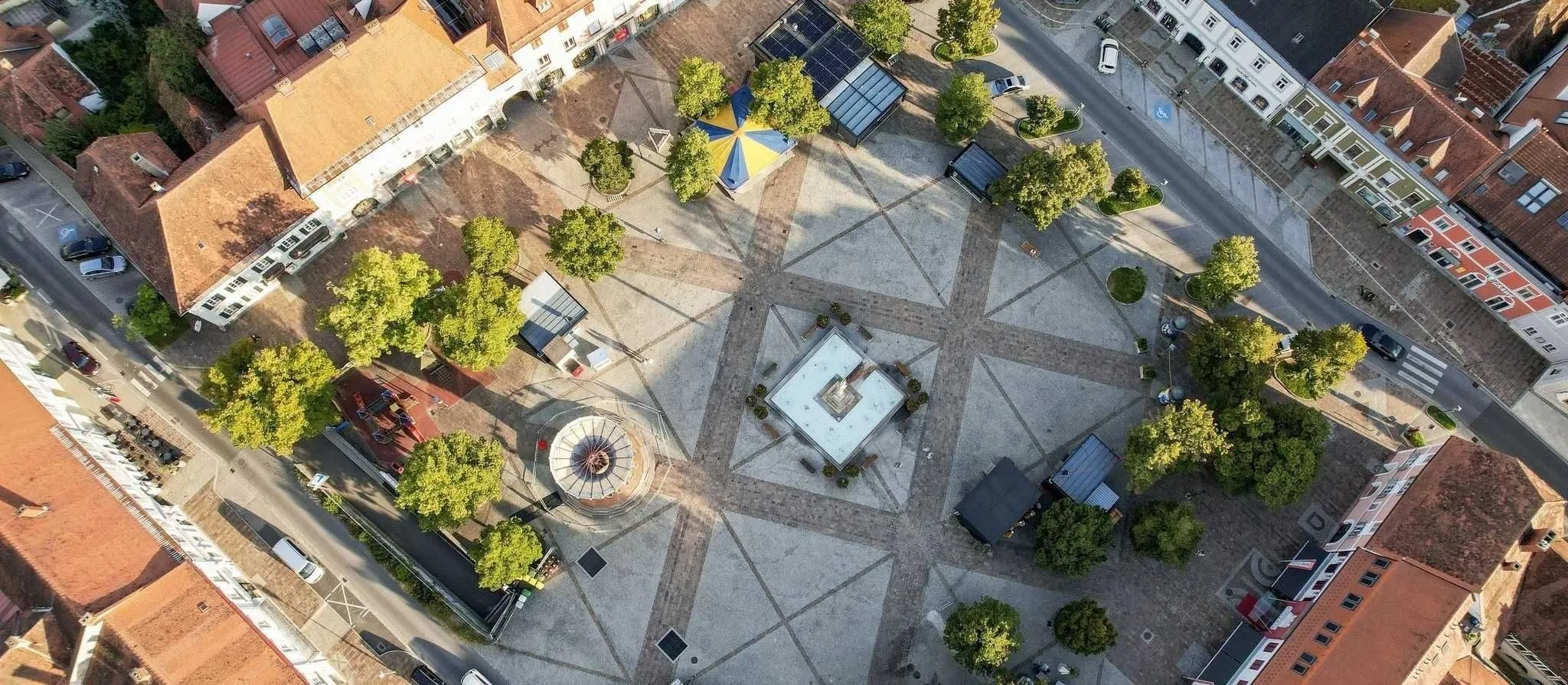 An aerial view of a city square with brick pavement, a fountain, trees, solar panels, and buildings. There are cars parked on the street and an umbrella tent.