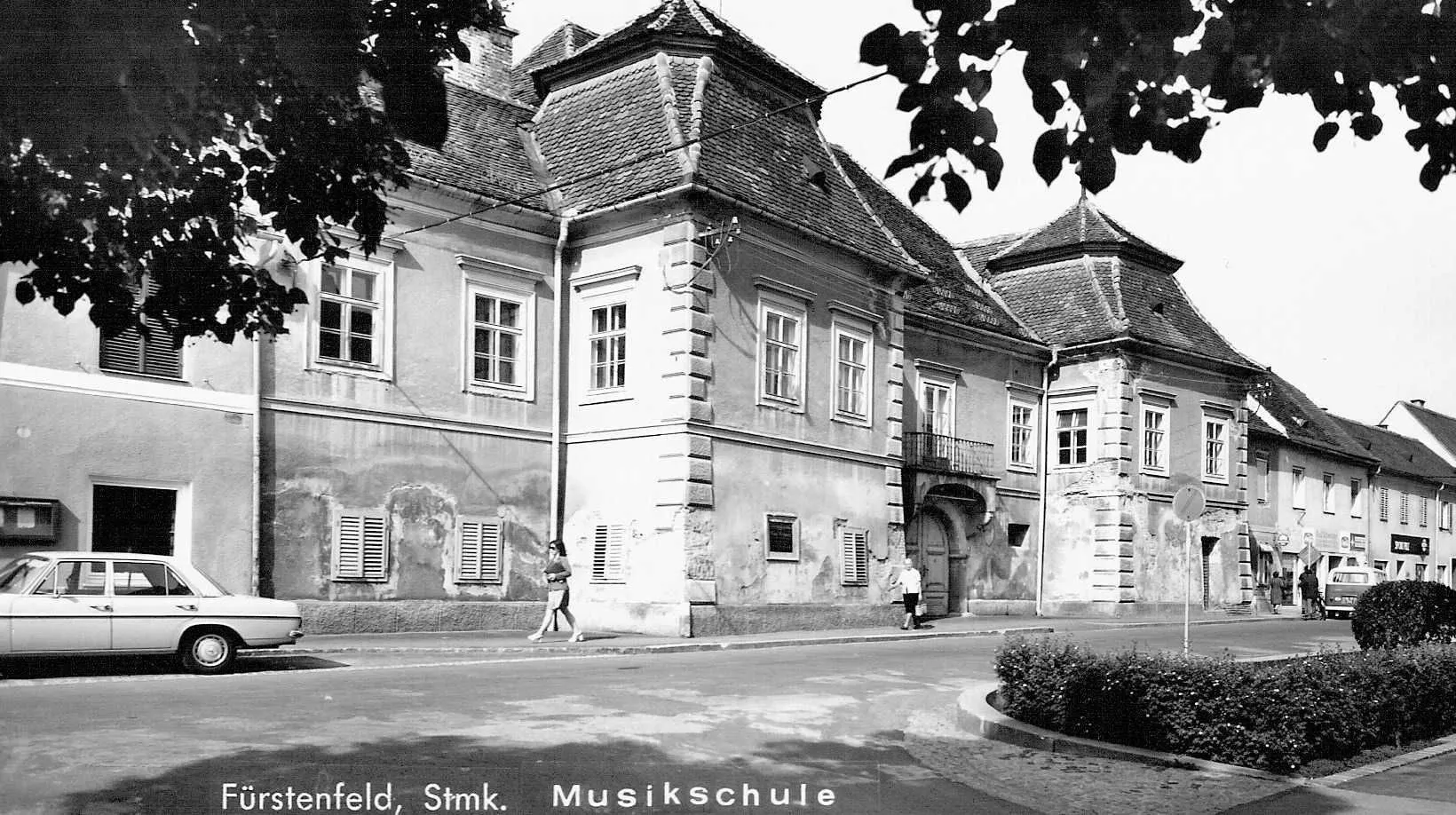 A black-and-white photo shows an old building with a tiled roof, many windows, and two people walking on the sidewalk. The building has a balcony, and the word 'Musikschule' is written at the bottom.