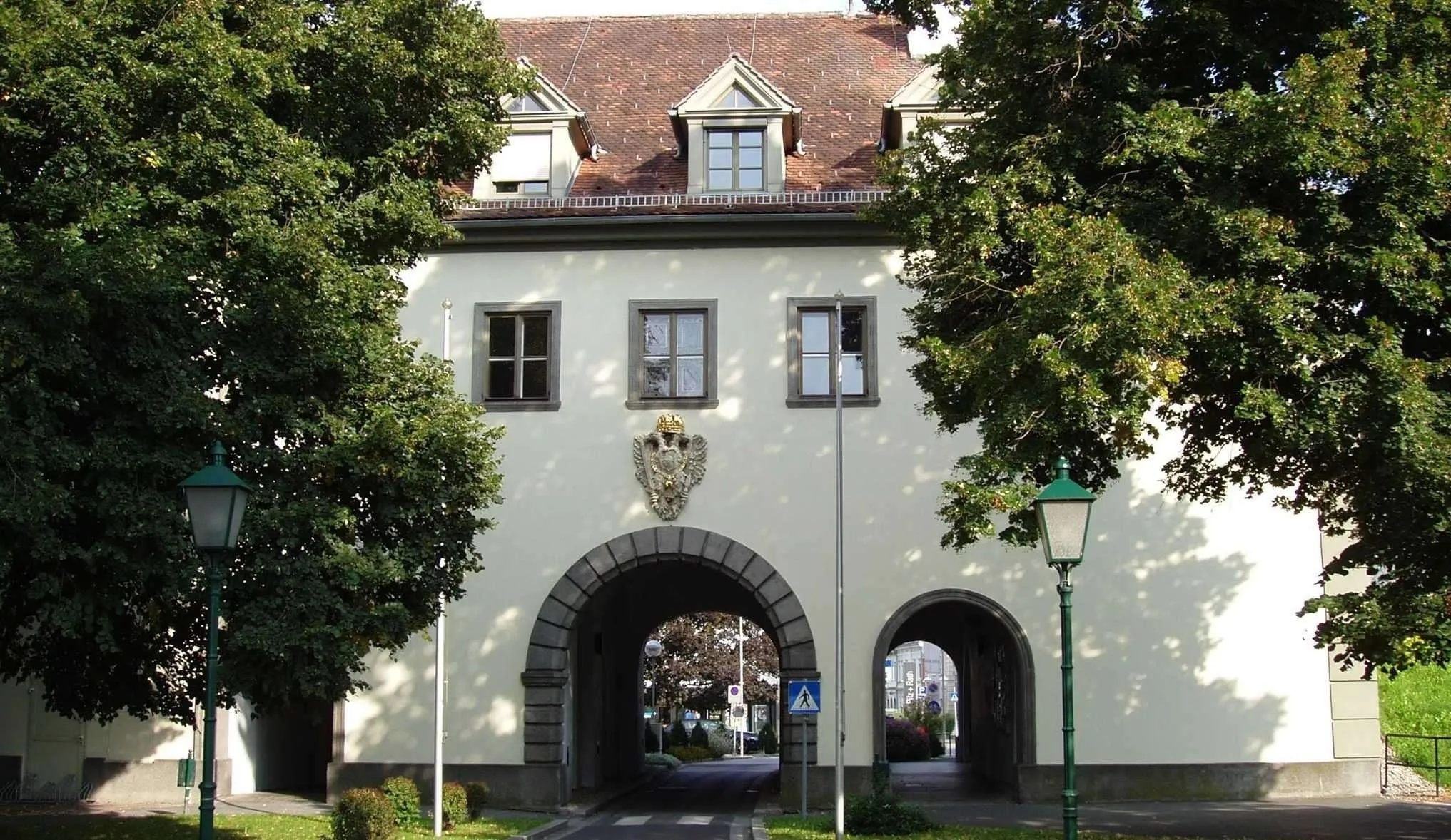 A traditional German building with a brown roof and arched entryways. A coat of arms is above the middle window. Trees and a streetlight line the path.