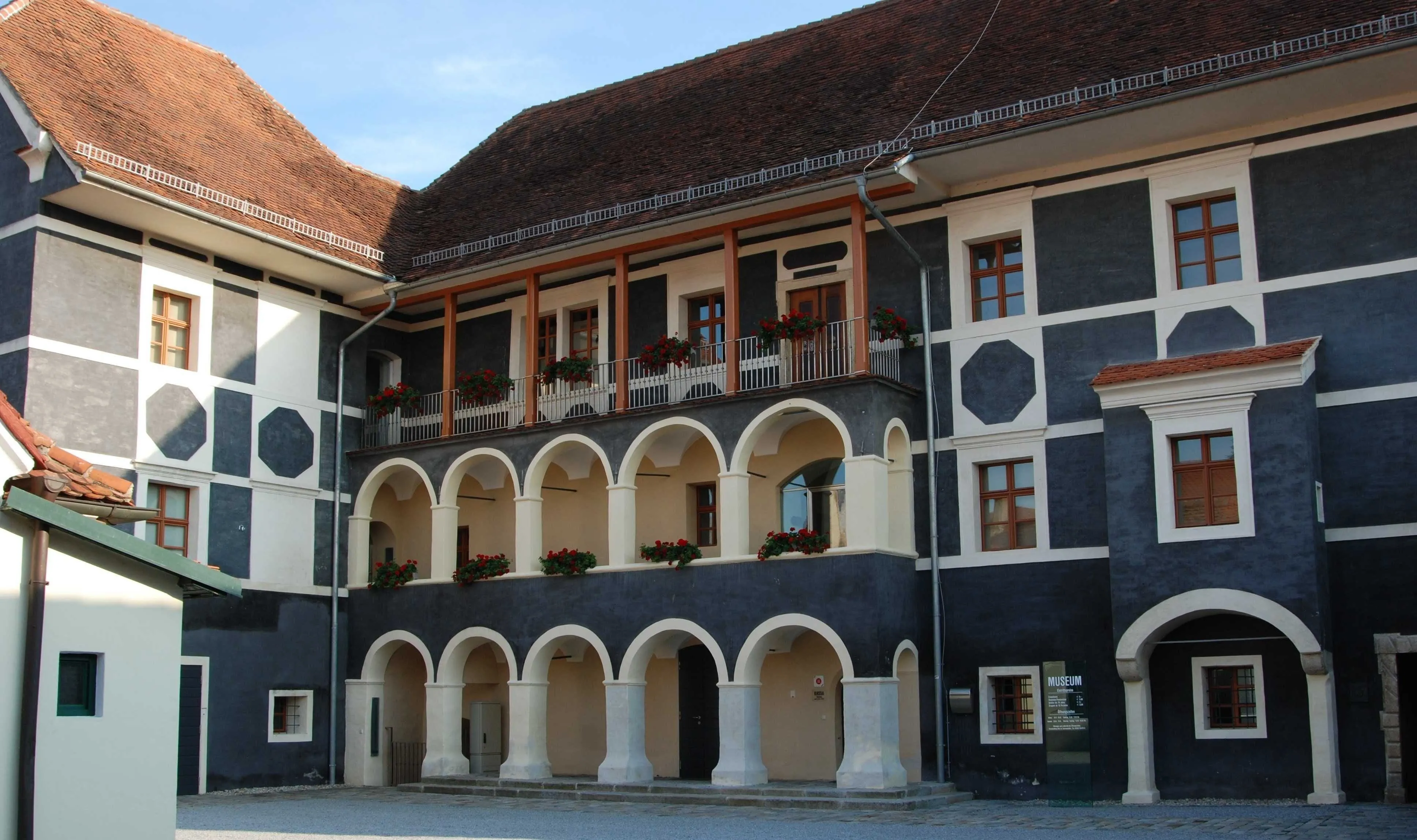 A historical building with black and white exterior walls features multiple arched windows and a balcony adorned with red flowers. The main entrance is flanked by pillars, and a sign on the right side reads 'Museum'.