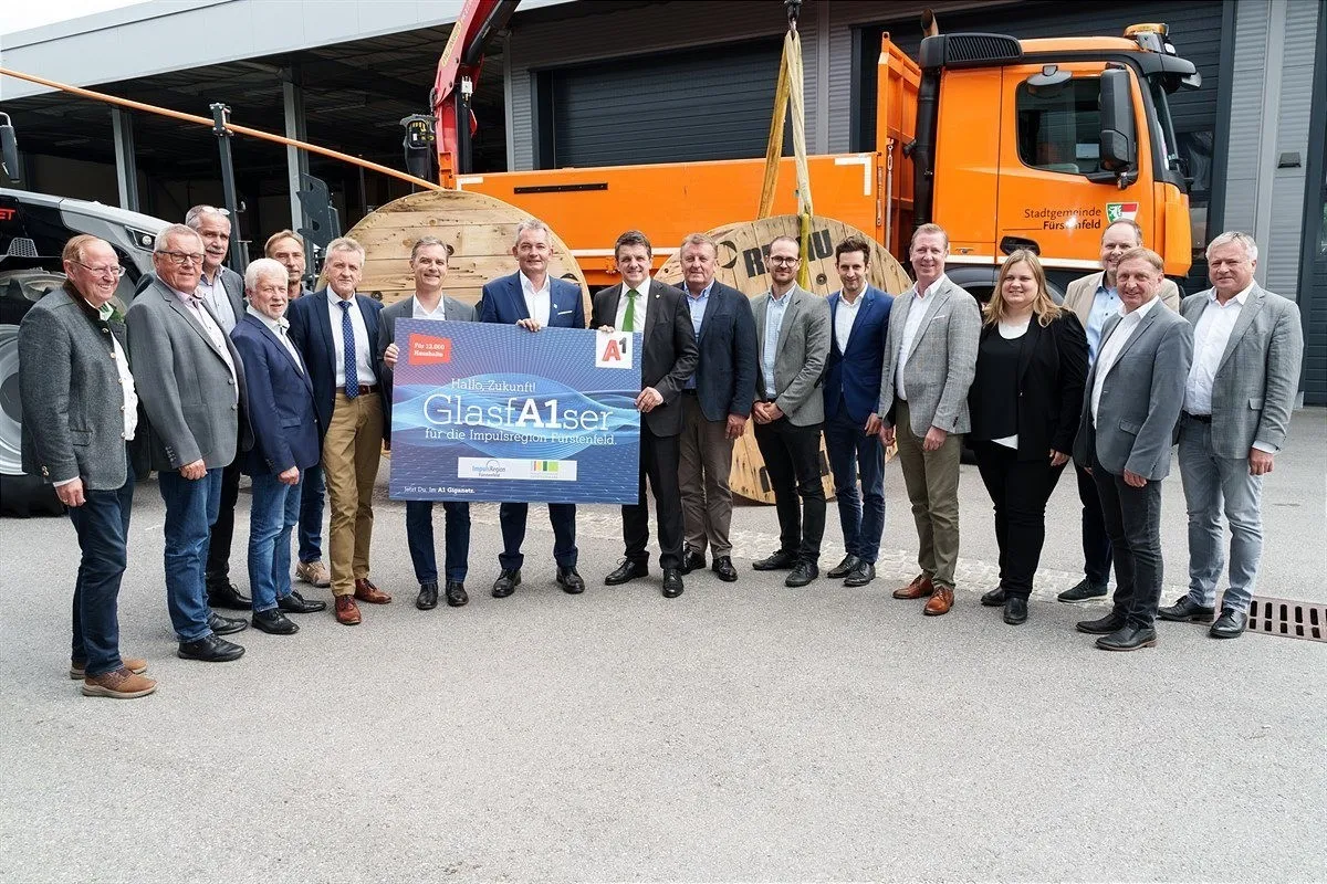 A group of people in suits and ties are standing next to each other in front of a building. They are holding a large certificate that reads 'GlasA1ser'. Behind them, a crane is lifting a large cable spool.