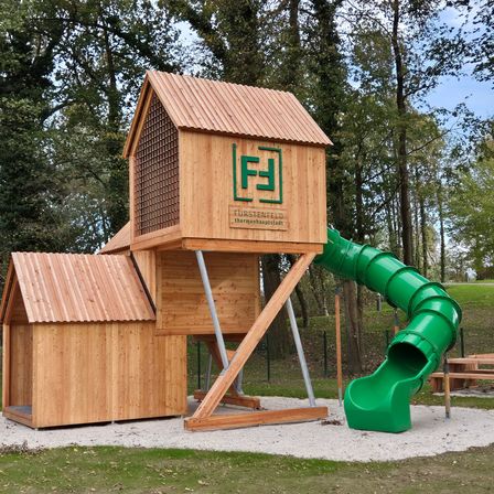 Ein hölzerner Spielplatz mit einer grünen Rutsche und einer Bank befindet sich in einem grasigen Bereich, umgeben von Bäumen.