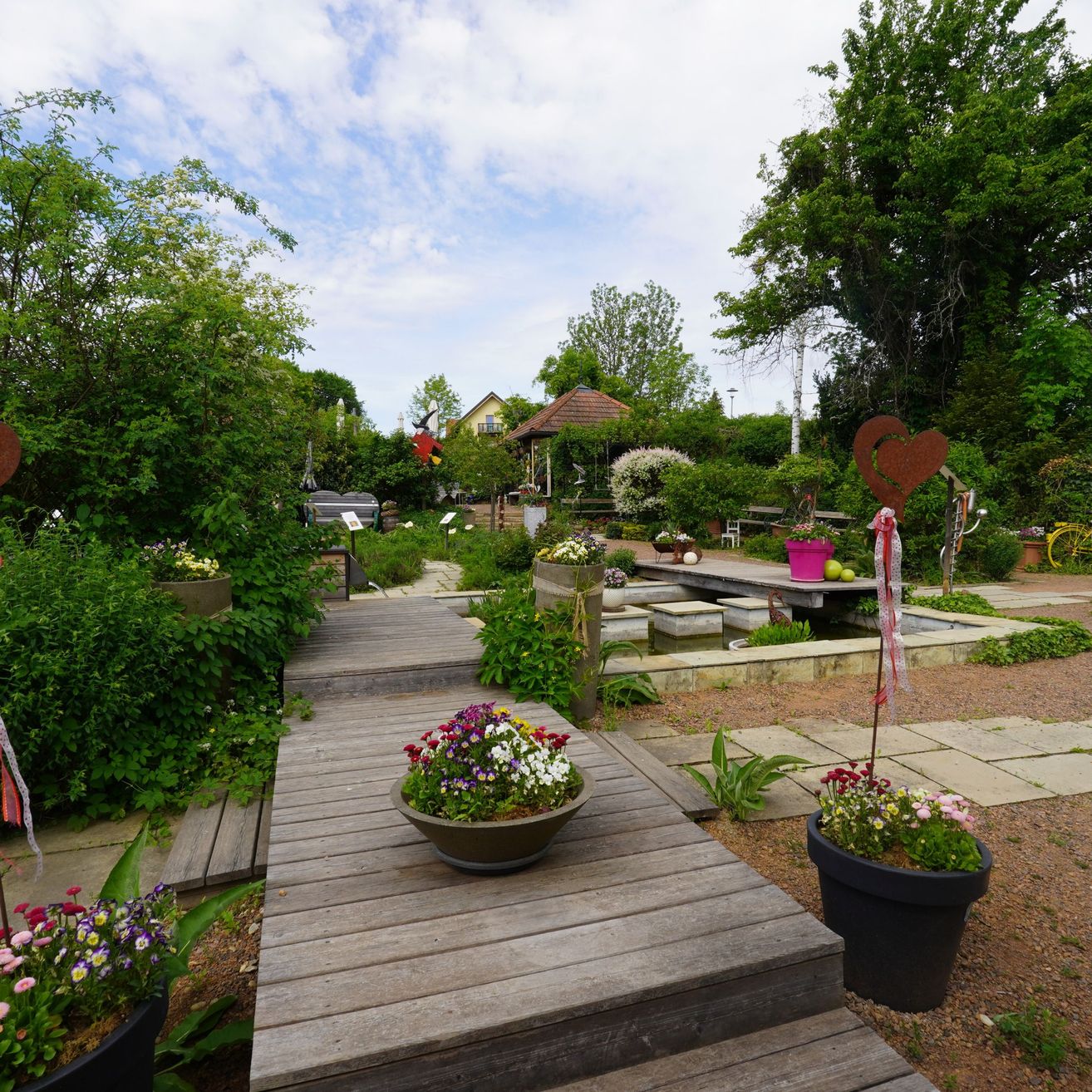 A pathway made of wood leads through a garden filled with flowers, plants, and trees.