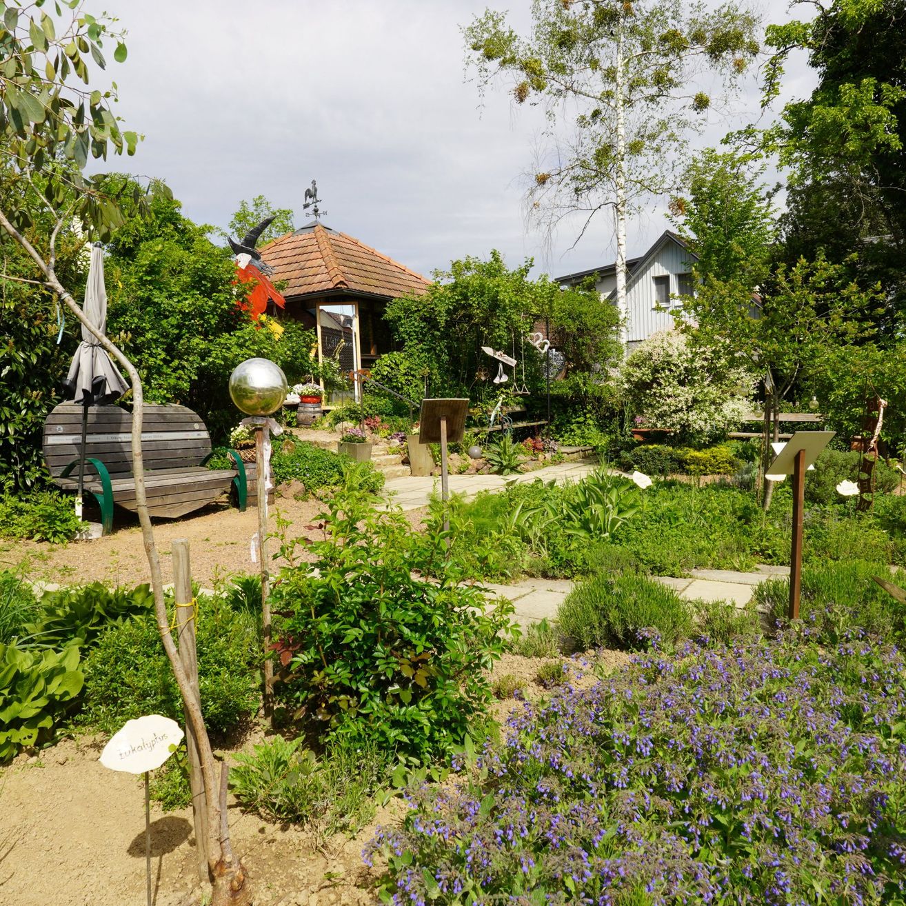 An outdoor area with a bench, a metal globe, and purple flowers. Trees and shrubs surround the area, and there are houses and a gazebo in the distance.