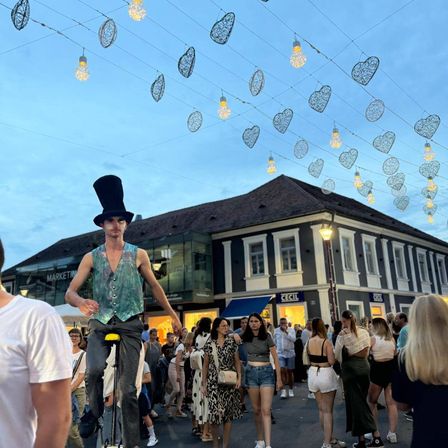 Ein Straßenkünstler reitet auf einem Einrad auf einer überfüllten Straße mit dekorativen Lichtern. Die Menge beobachtet ihn, wie er auf dem Einrad balanciert. Die Straße ist gesäumt von Gebäuden und Menschen.