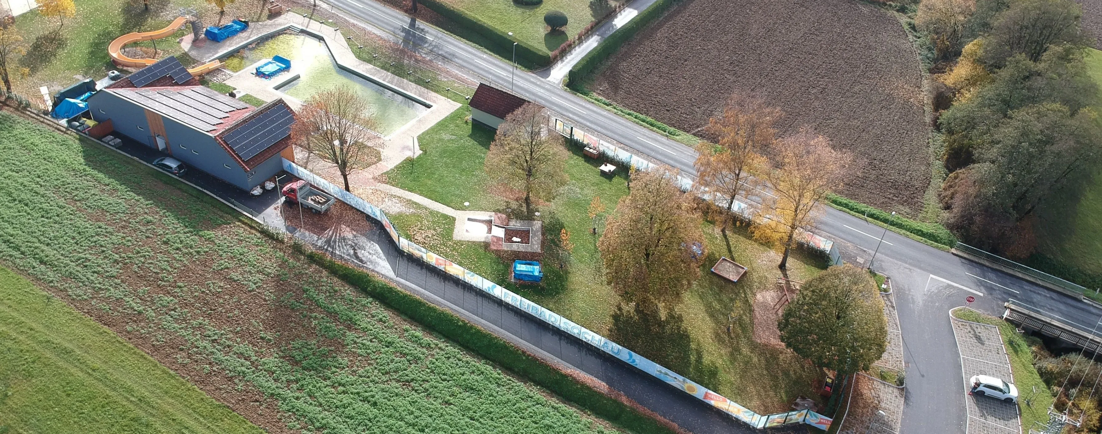 Eine Vogelperspektive einer Landschaft mit einem kleinen Haus, einer Straße und einem Feld. Bäume und ein blaues Objekt sind verstreut. Ein Zaun verläuft entlang der Seite der Straße.