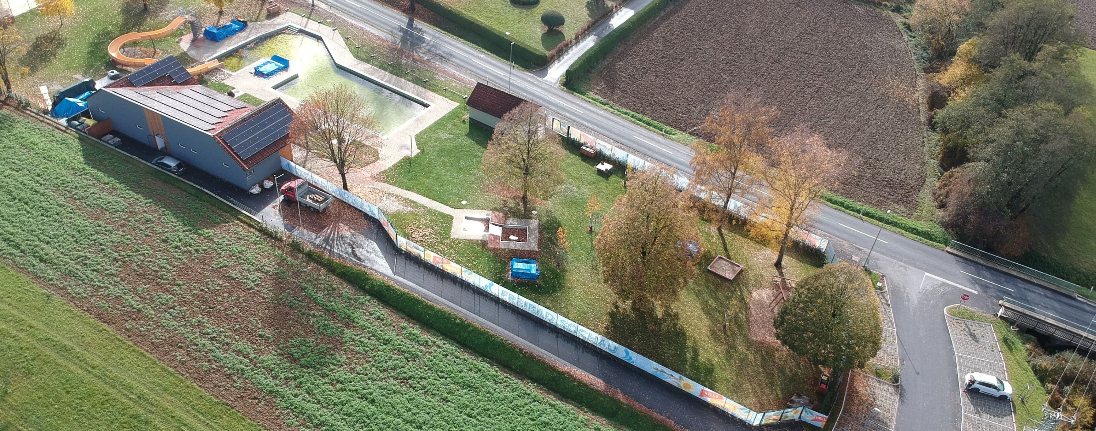 Eine Vogelperspektive einer Landschaft mit einem kleinen Haus, einer Straße und einem Feld. Bäume und ein blaues Objekt sind verstreut. Ein Zaun verläuft entlang der Seite der Straße.
