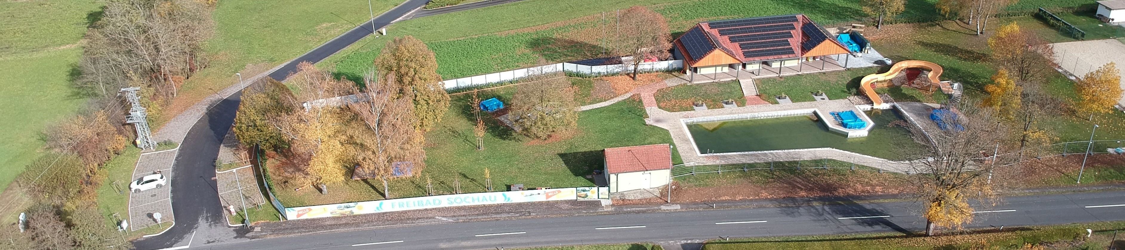 Eine Luftaufnahme einer Landschaft mit einem grünen Feld, einem kleinen Gebäude und einem abgedeckten Objekt in der Ecke. Bäume und Pflanzen sind verstreut.