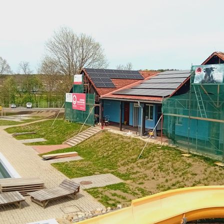 An aerial view of a building under construction with solar panels on the roof, surrounded by a green lawn, with a yellow slide and wooden deck chairs in the foreground.