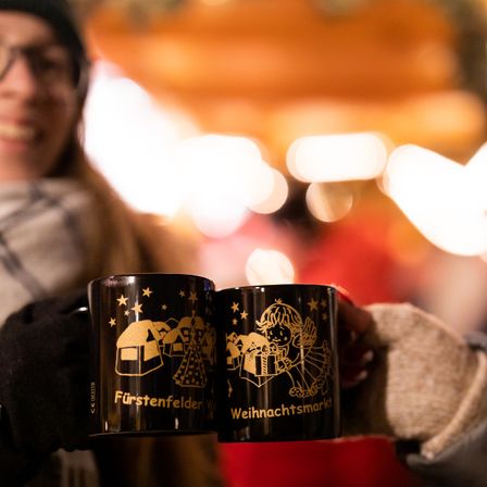 A woman holds two mugs with the words Furstenfelder and Weihnachtsmarkt. She smiles at the camera. Behind her, there are blurry lights and decorations.