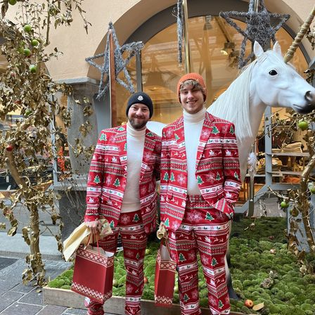 Two men wearing red Christmas sweaters and pants are standing in front of a white horse statue. They are holding red gift bags. Behind them is a Christmas tree with ornaments and a glass window with shelves inside.