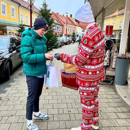A woman in a green jacket holds a bag and hands it to a man in Christmas pajamas who holds a red gift bag on a street with parked cars and buildings.