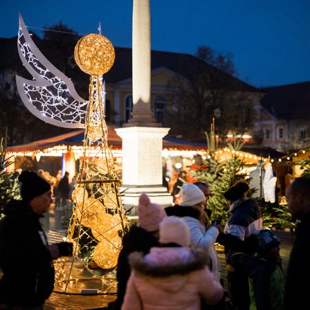 A family of three stands in front of a Christmas tree with decorative lights. Behind them, a city square with more Christmas trees and festive decorations. In the distance, buildings are illuminated. The sky is dark and cloudy.