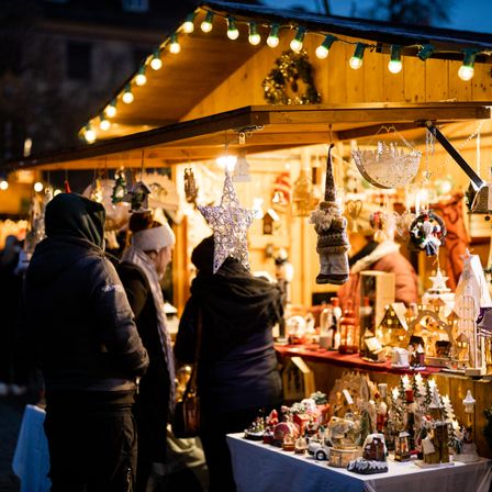 Mehrere Personen stehen vor einem Marktstand, der mit Weihnachtslichtern und Dekorationen geschmückt ist.