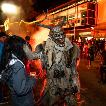 Eine als Dämon verkleidete Person bläst Feuer aus seinem Mund vor einer Menge. Eine Frau beugt sich vor, um ihn zu küssen.
