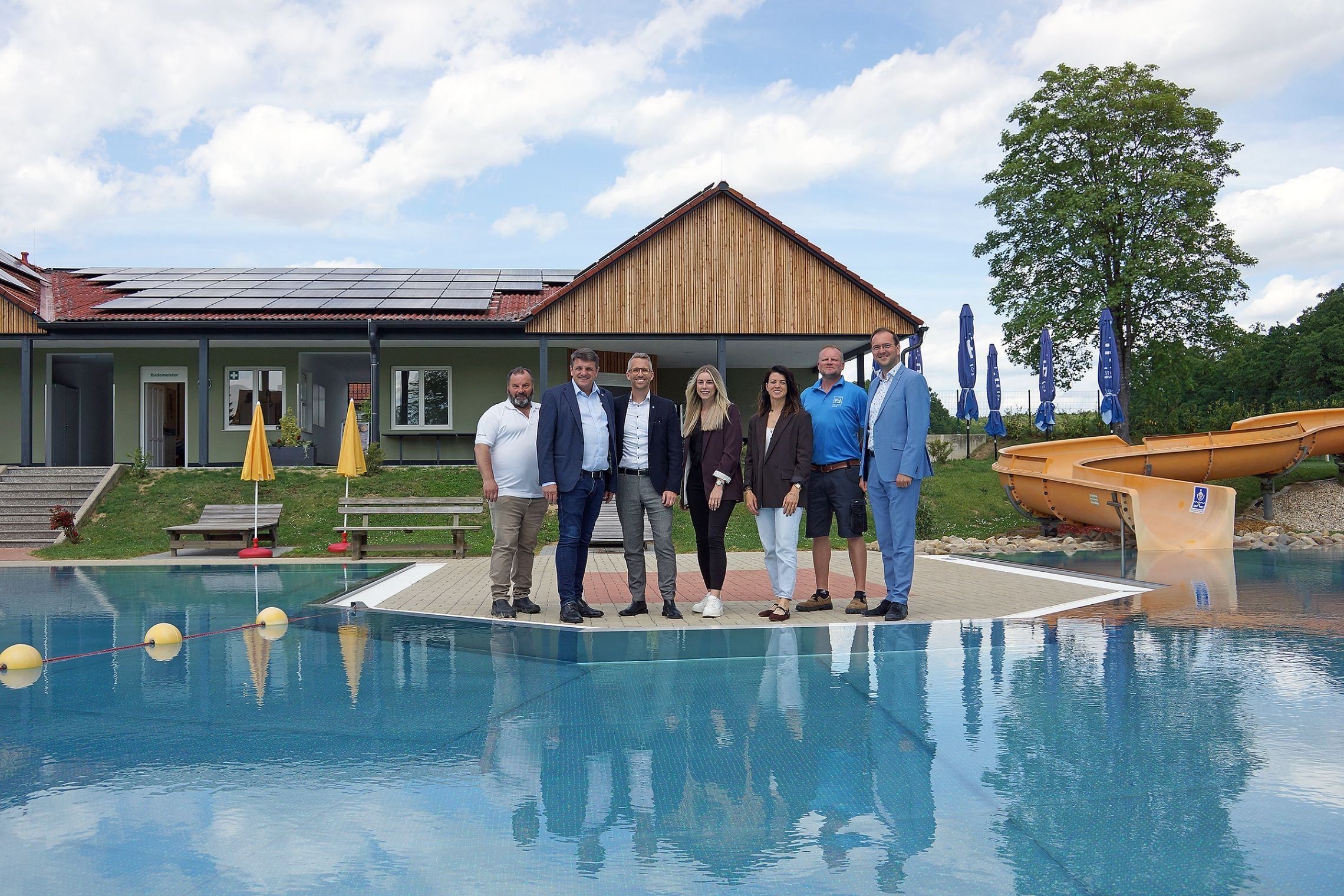 Eine Gruppe von Menschen steht vor einem Haus mit Solarpanelen, in der Nähe eines Schwimmbeckens. Das Haus hat eine Holzfassade und ein rotes Dach. Sie posieren für ein Foto an einem sonnigen Tag.