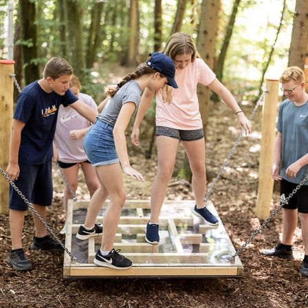Eine Gruppe von Kindern spielt ein Spiel mit Sprungsteinen in einem Waldgebiet, wobei zwei Mädchen auf den Sprungsteinen stehen.