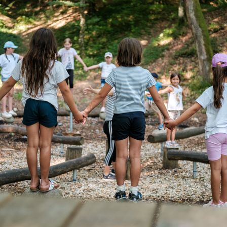 Eine Gruppe von Kindern spielt auf dem Spielplatz. Sie halten sich an den Händen und gehen auf den Holzhindernissen.