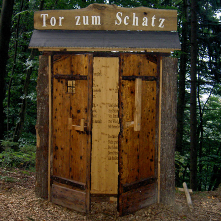 Ein hölzernes Häuschen im Wald mit einem Schild, das 'Tor zum Schatz' besagt. Das Häuschen hat zwei Holztüren mit einem Fenster in der Mitte.