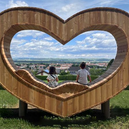 Zwei Kinder sitzen in einer holzherzförmigen Bank und blicken auf eine kleine Stadt.
