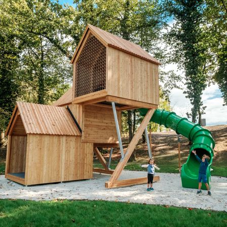 Zwei Kinder spielen an einem hölzernen Spielhaus mit einer grünen Rutsche in einem Waldgebiet.